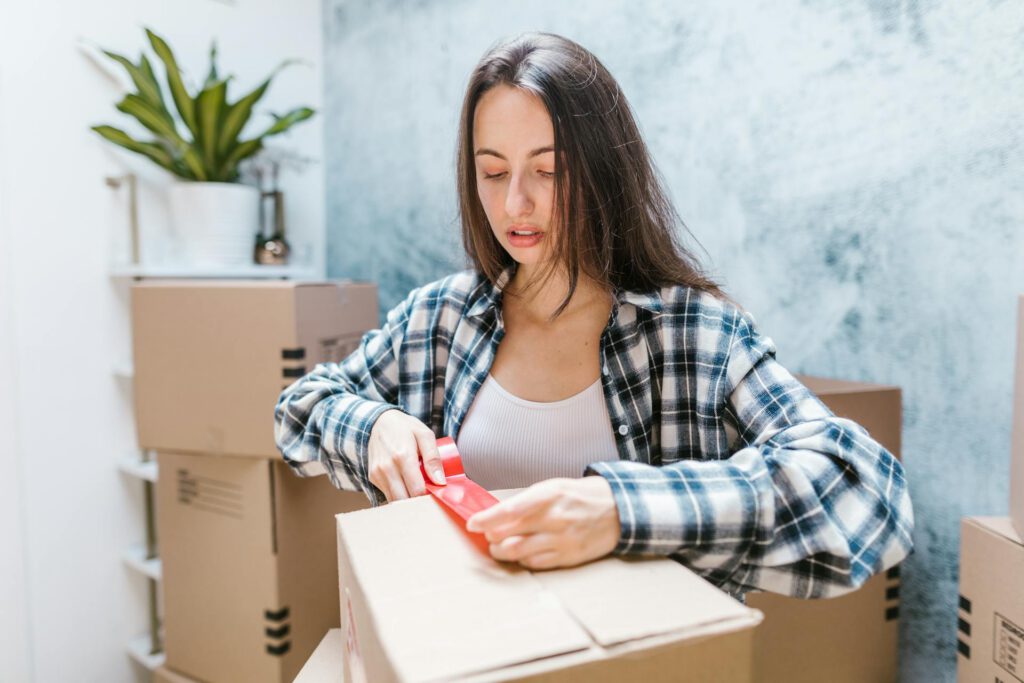 A woman sealing a cardboard box with red tape, surrounded by boxes.