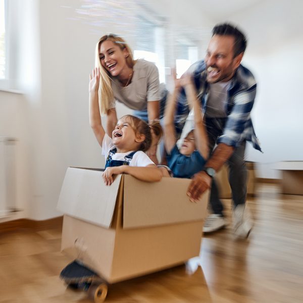 Family plays with cardboard box on skateboard in sunny living room.